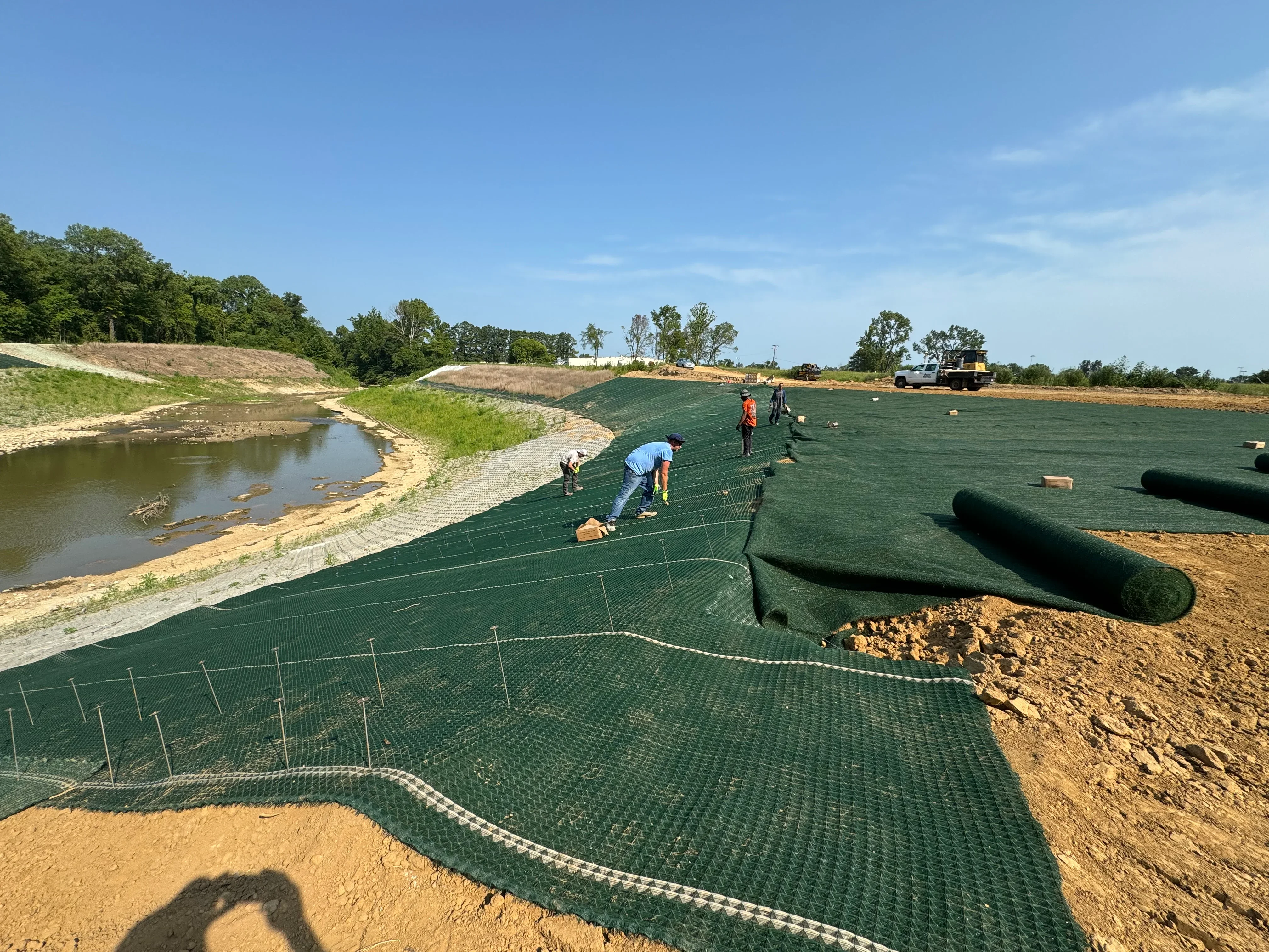 Green erosion control matting installed on sloped banks with stakes securing geotextile fabric along waterway