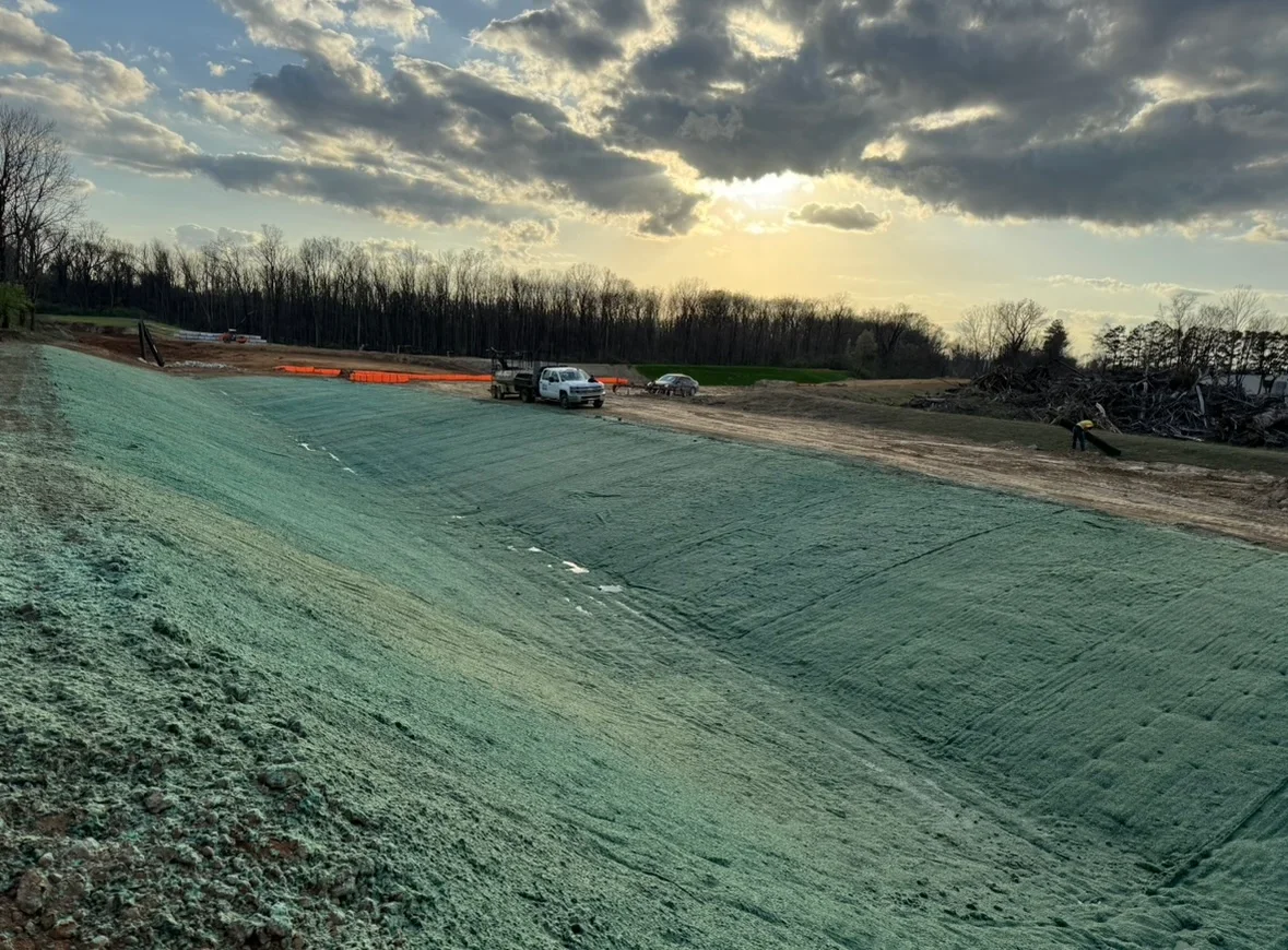 Green erosion control matting installed on prepared soil with orange fencing and construction vehicles