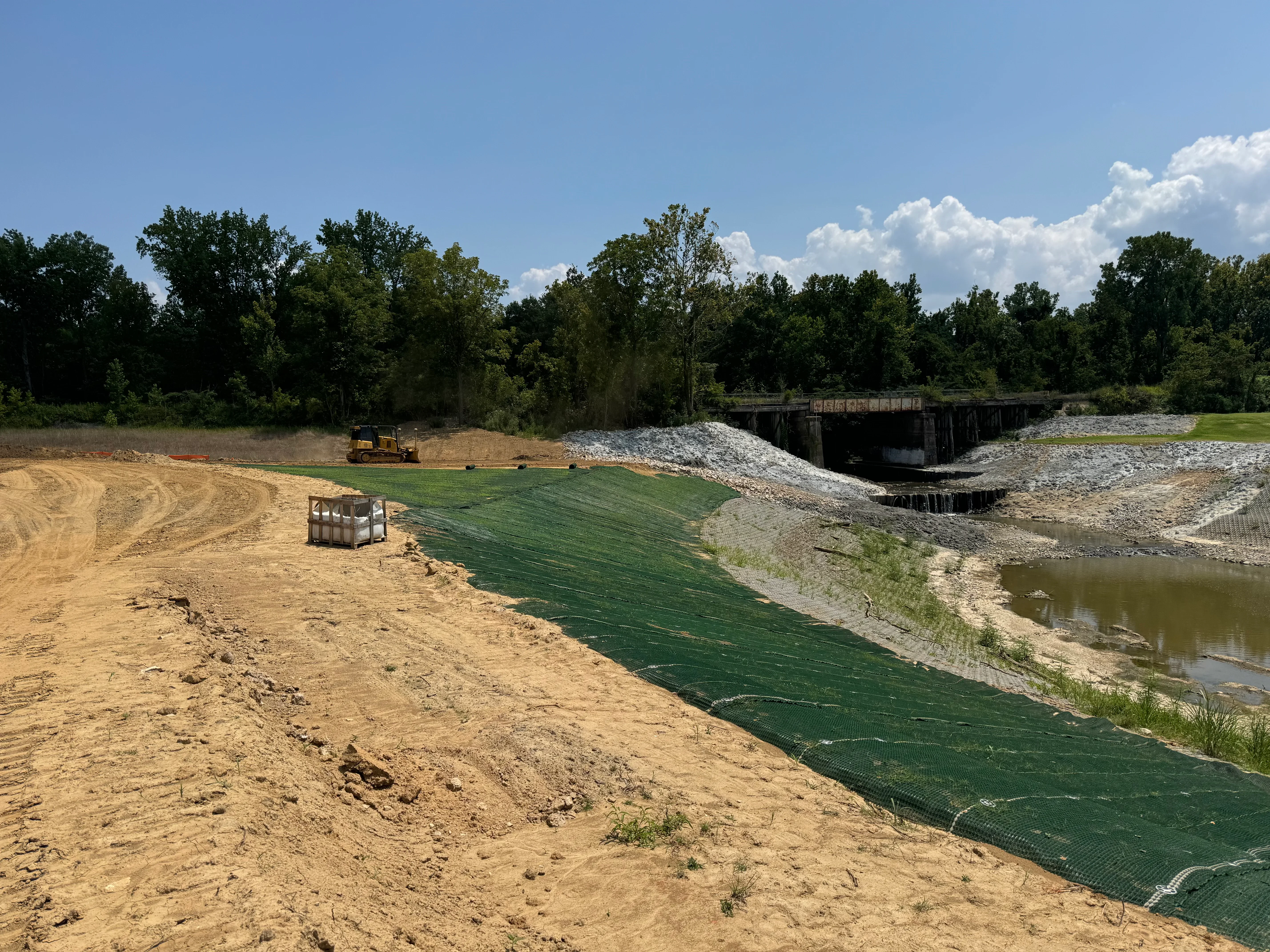 Green geotextile erosion control matting installed on riverbank slope with staged aggregate and grading equipment